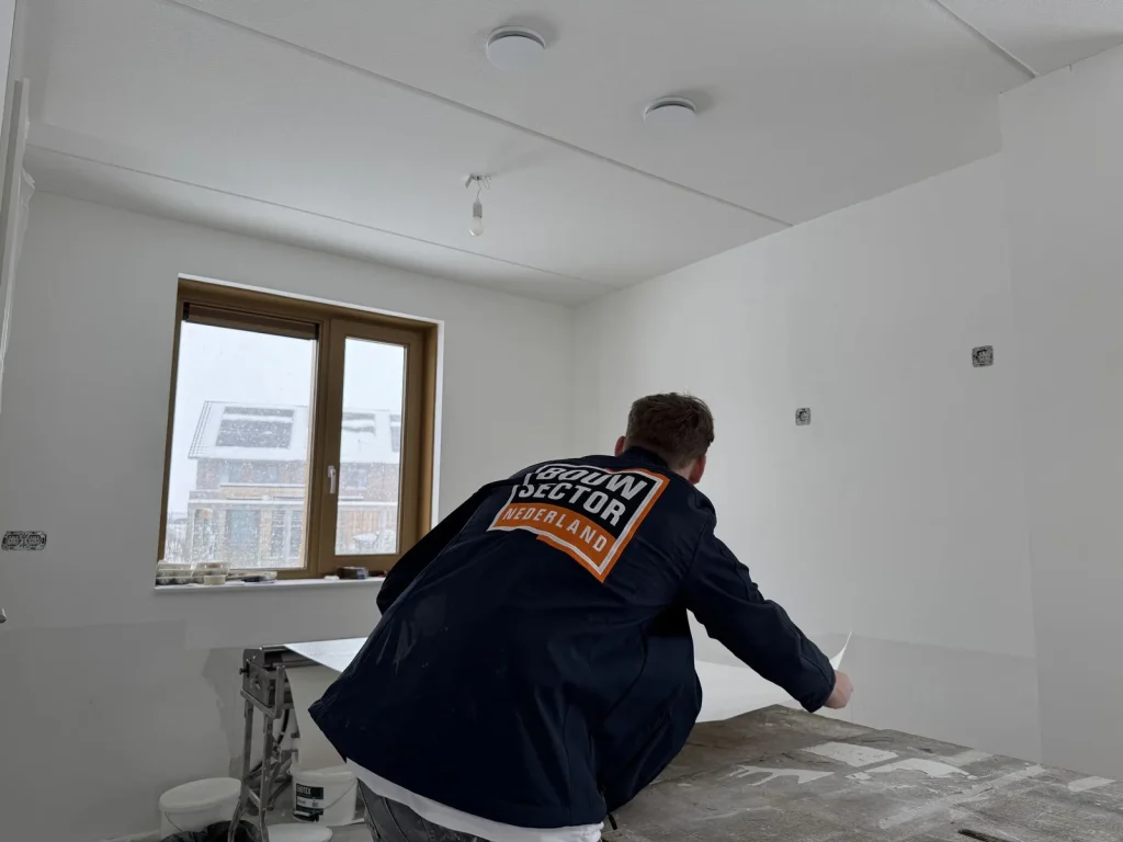 Behanger van Bouwsector Nederland rolt een baan vlies uit op de plaktafel in een kale kamer met sneeuw buiten