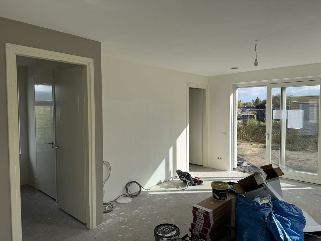 Woonkamer tijdens de verbouwing met dozen, verfemmers en kabels op de vloer en open deuren naar de gang