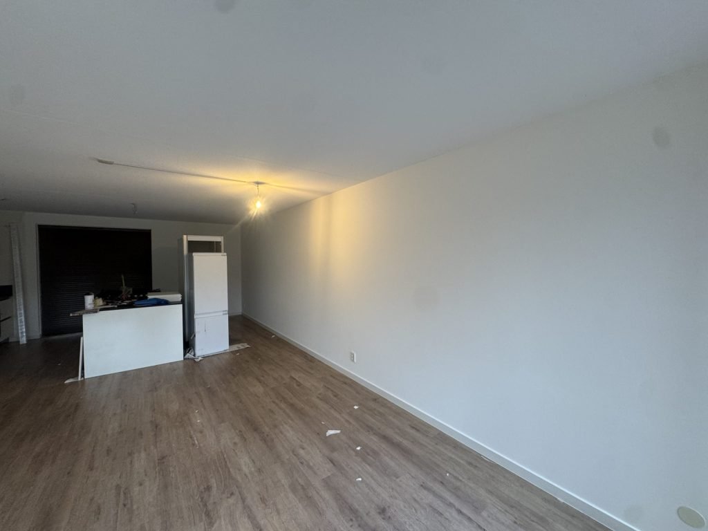 Lange woonkamer met strak afgewerkte witte wand, houten vloer en een keukenblok achterin