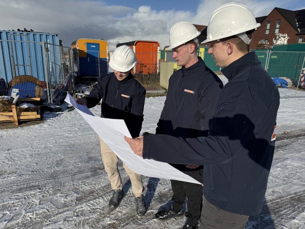 Drie vakmensen van Bouwsector Nederland met witte helm bekijken samen een werktekening op een bouwplaats