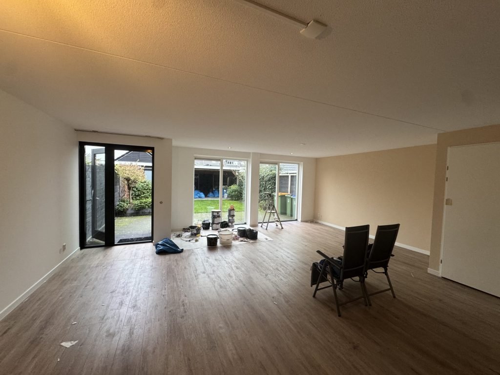 Lege woonkamer met tuindeuren tijdens een klus, met verfemmers en een trapje op afgedekte vloer